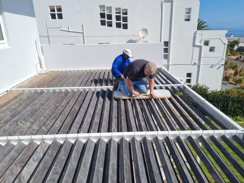 Two men are measuring and cutting wood planks on a rooftop deck with a view of a coastal area in the background.